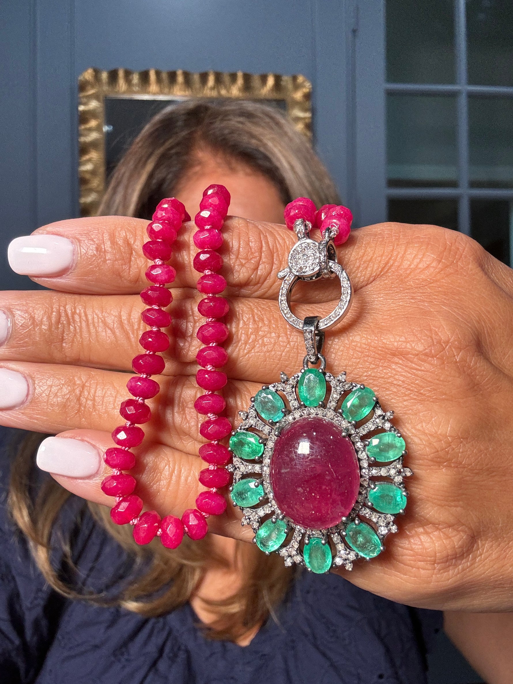 Hand displaying an 18-inch ruby bead necklace with ornate emerald halo pendant featuring central red stone and diamond-accented sterling silver clasp