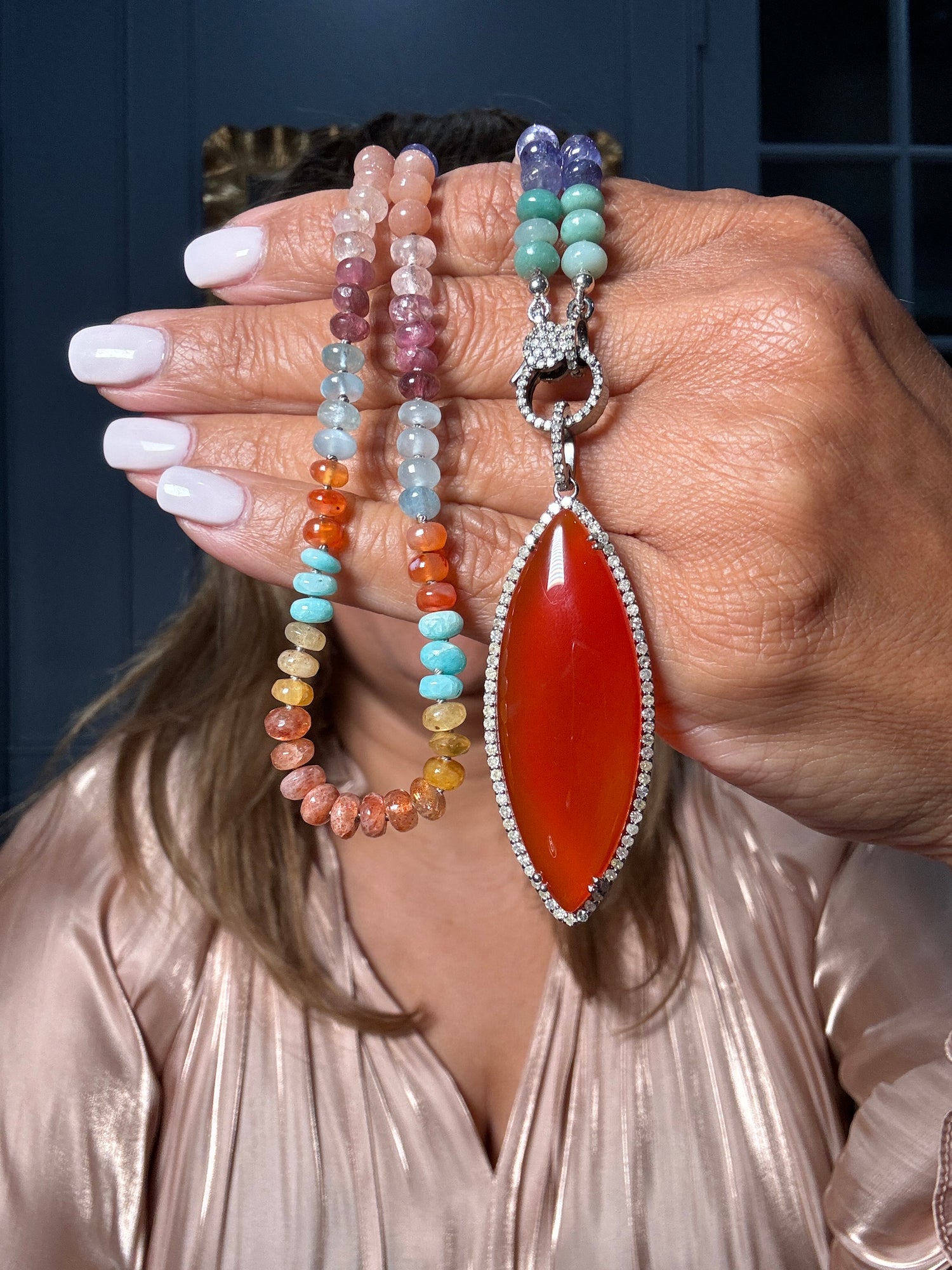 Hand holding 21.5-inch multicolor gemstone necklace featuring carnelian, aquamarine, tourmaline, and other natural stones with diamond-encrusted sterling silver clasp and large orange carnelian pendant.