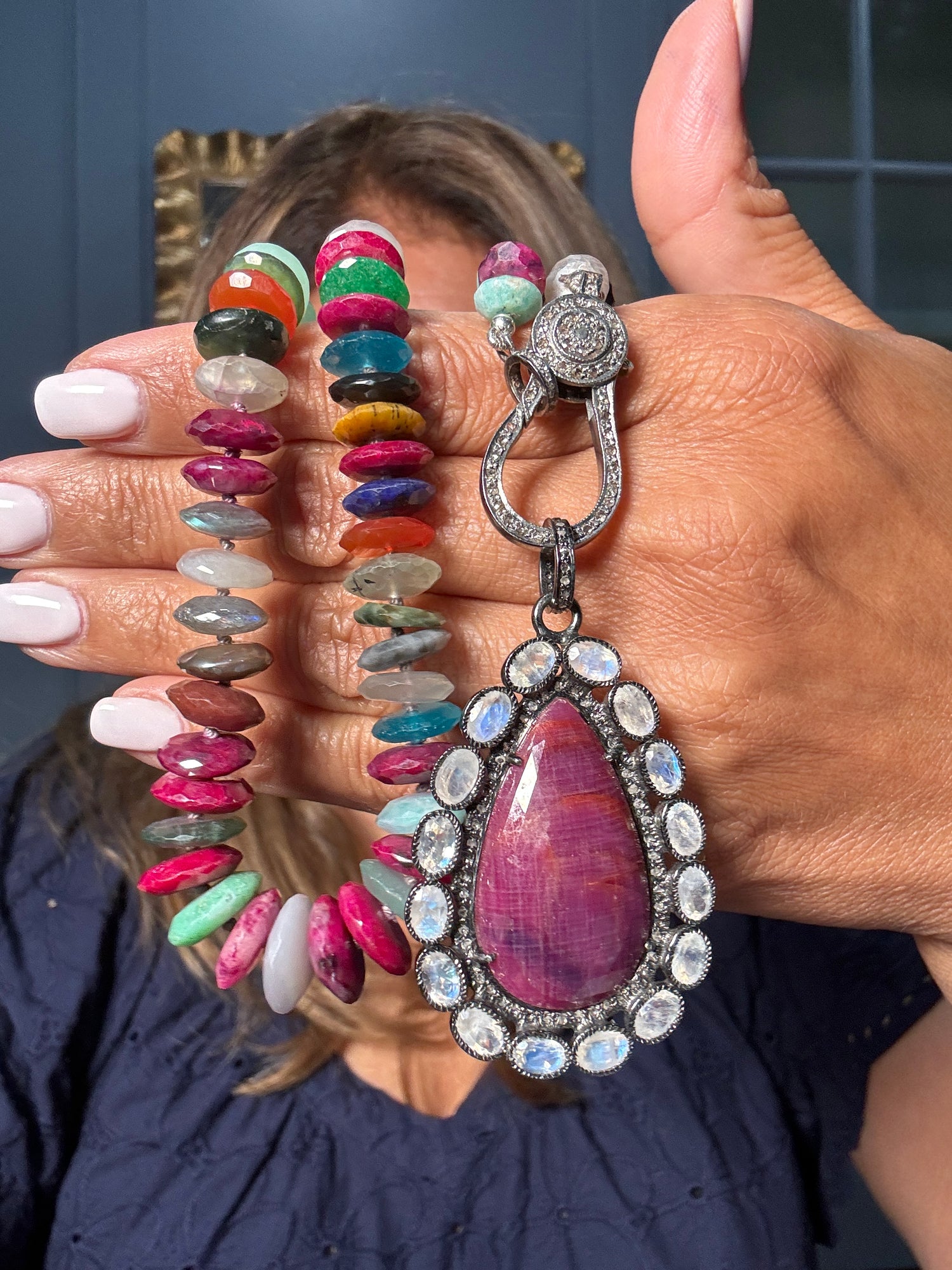 Colorful 22-inch mixed gemstone necklace with oval-cut ruby, emerald, sapphire, and agate beads, featuring teardrop ruby moonstone pendant with diamond halo clasp, held in cupped hands