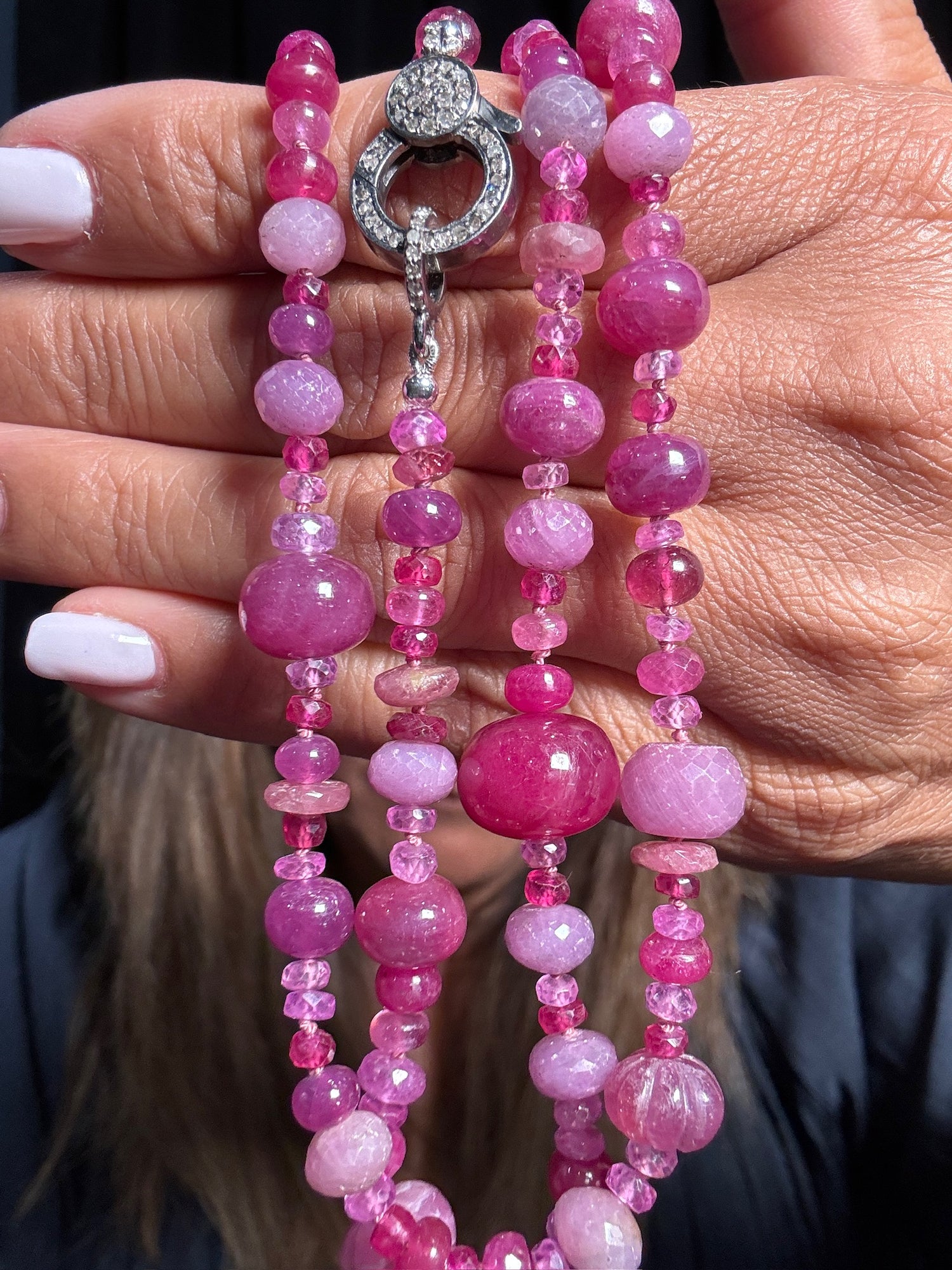 Hand holding 26-inch ruby and pink sapphire beaded necklace with diamond-encrusted sterling silver clasp featuring graduated oval and round gemstones