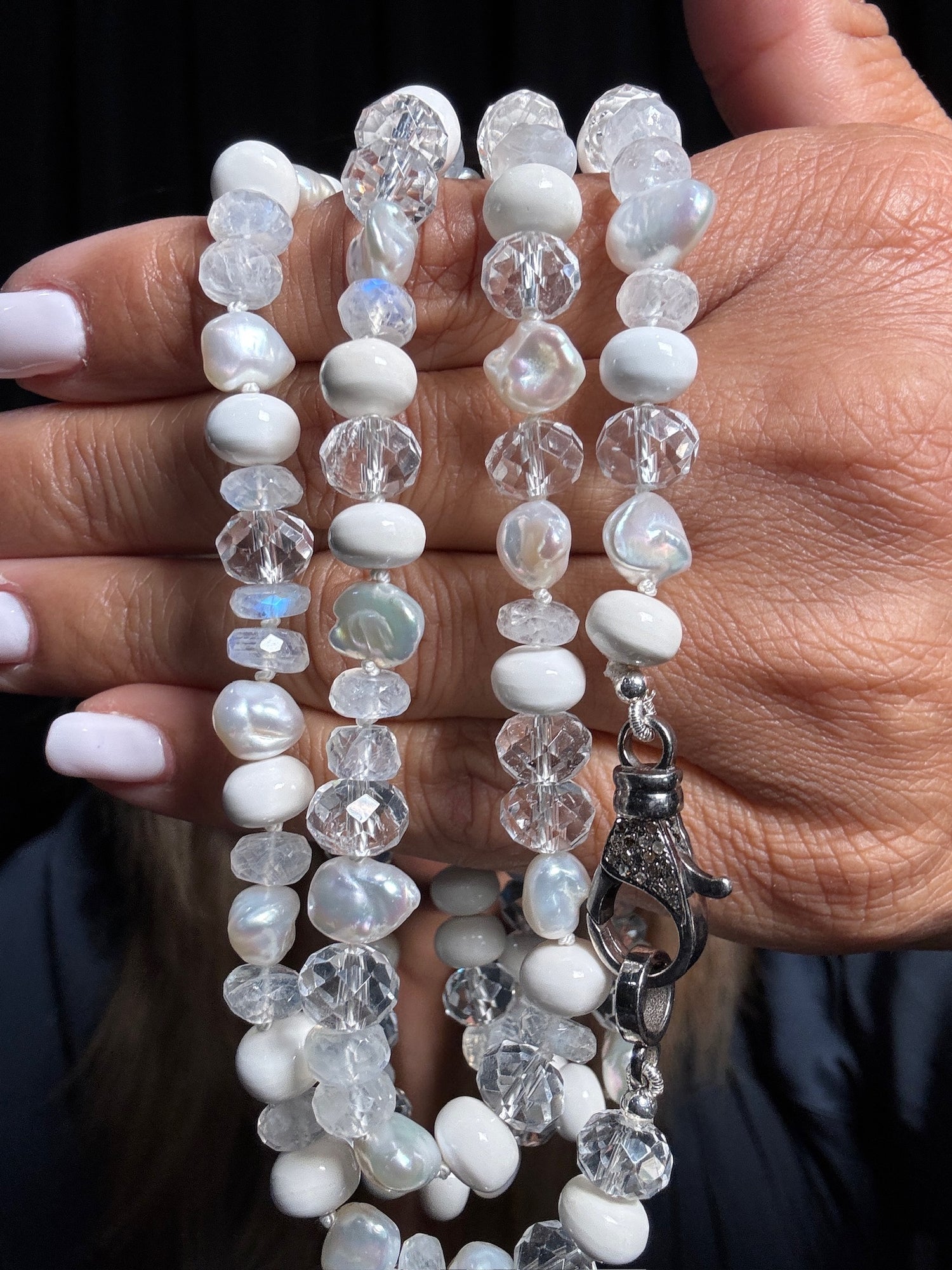 Hand holds 36-inch white gemstone necklace featuring moonstone, clear quartz, white pearls and agate beads with sterling silver lobster clasp against black background