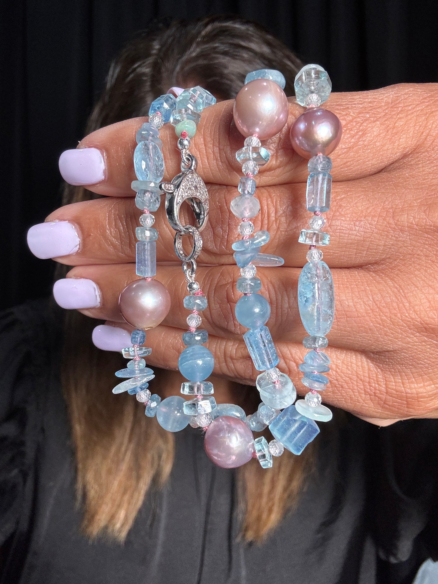 Hand holding a 21-inch aquamarine and pink Edison pearl necklace with various blue gemstone shapes and sterling silver clasp against dark background
