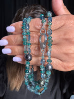 Hand holding 36-inch grandidierite and Tahitian pearl necklace with teal blue gemstones, dark gray pearls, and sterling silver diamond clasp against blonde hair background
