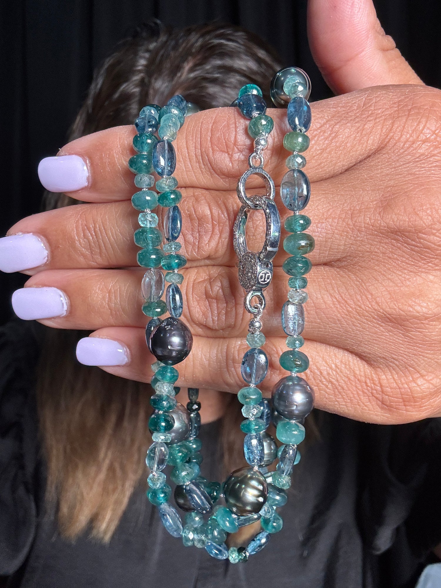Hand holding 36-inch grandidierite and Tahitian pearl necklace with teal blue gemstones, dark gray pearls, and sterling silver diamond clasp against blonde hair background