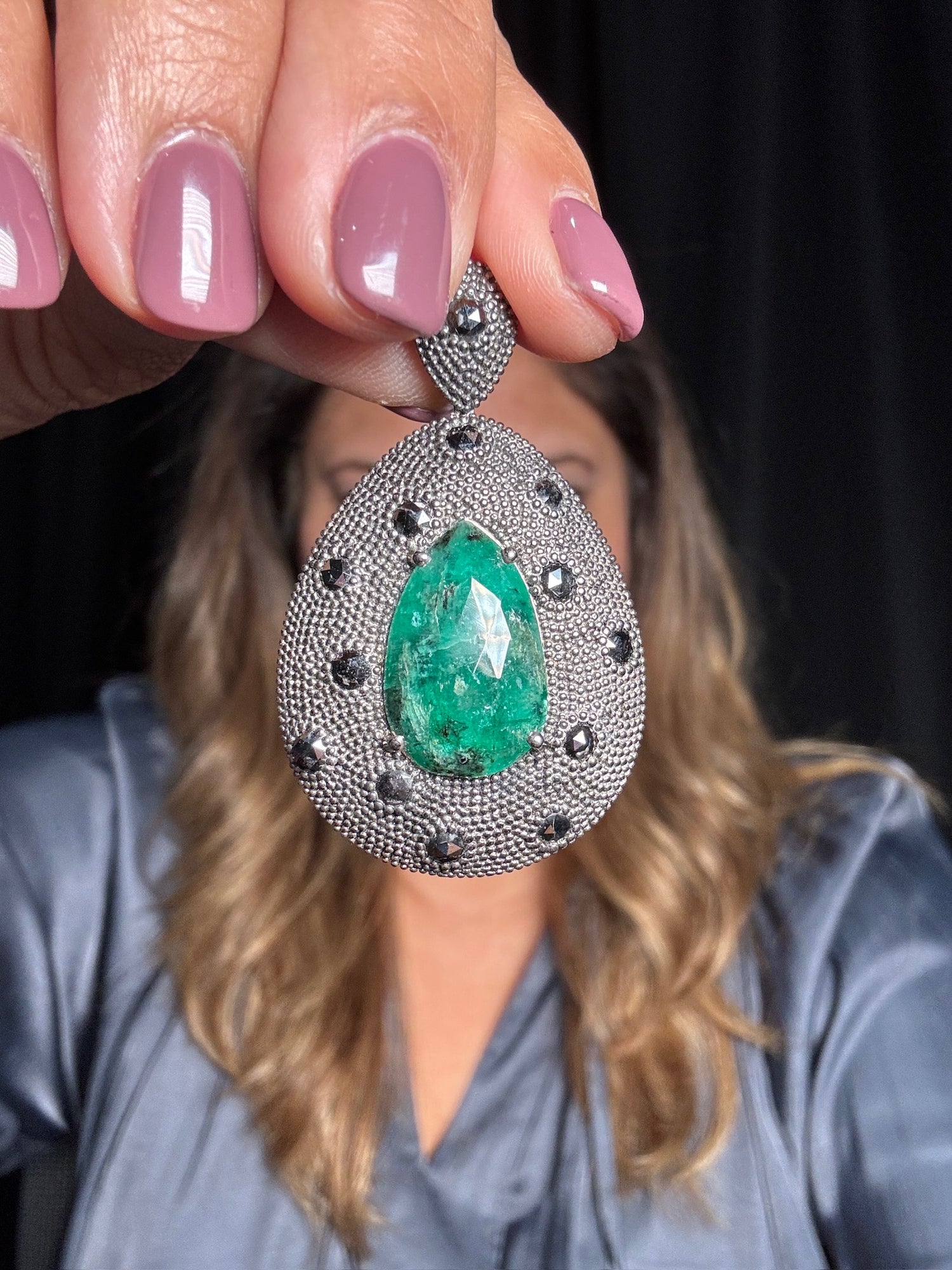 Hand holding large teardrop emerald pendant with diamond halo and black diamond accents in sterling silver against woman in blue shirt