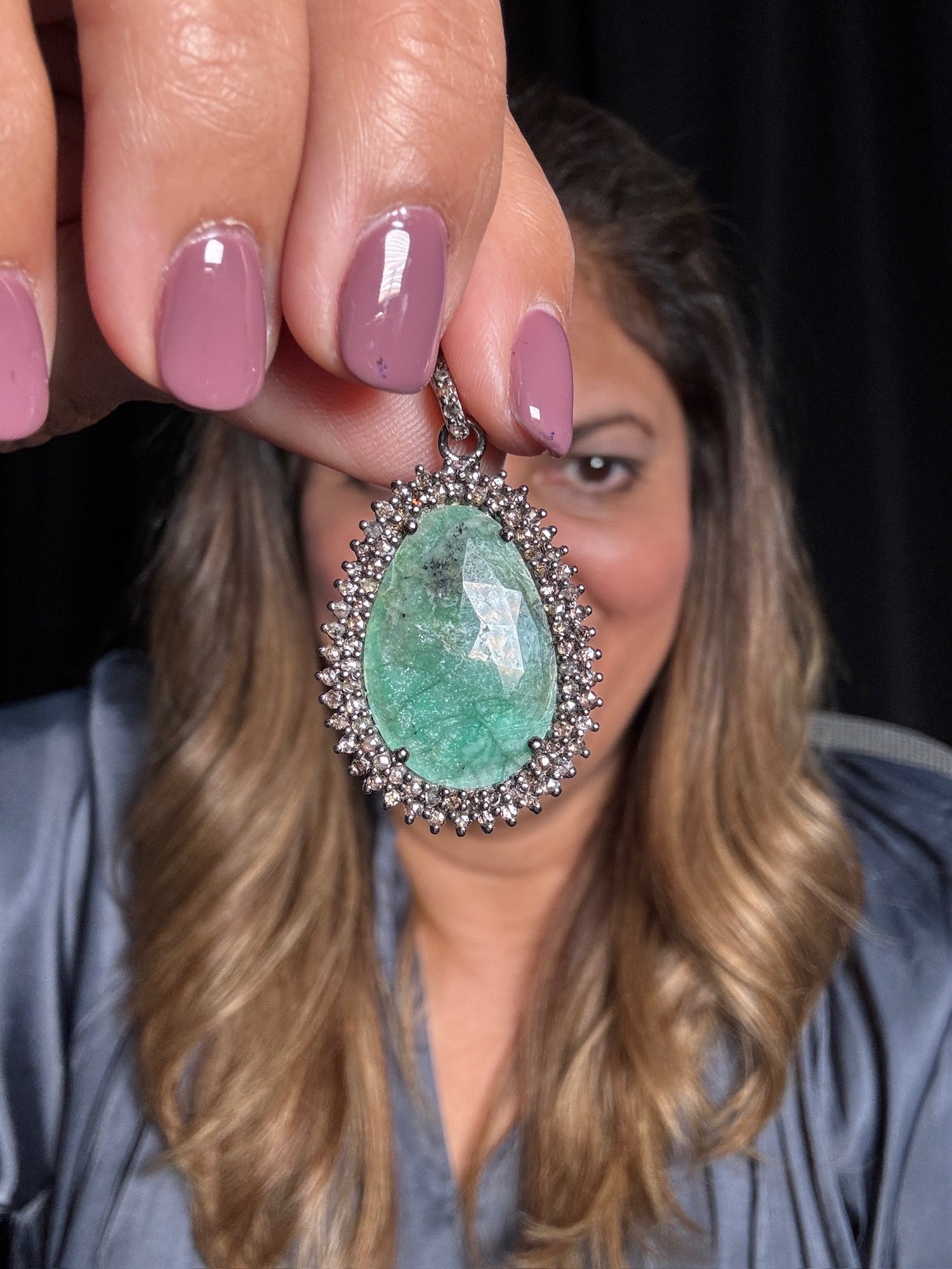 Hand holding large oval emerald teardrop pendant with sparkling diamond halo setting in sterling silver, woman with wavy blonde hair in background