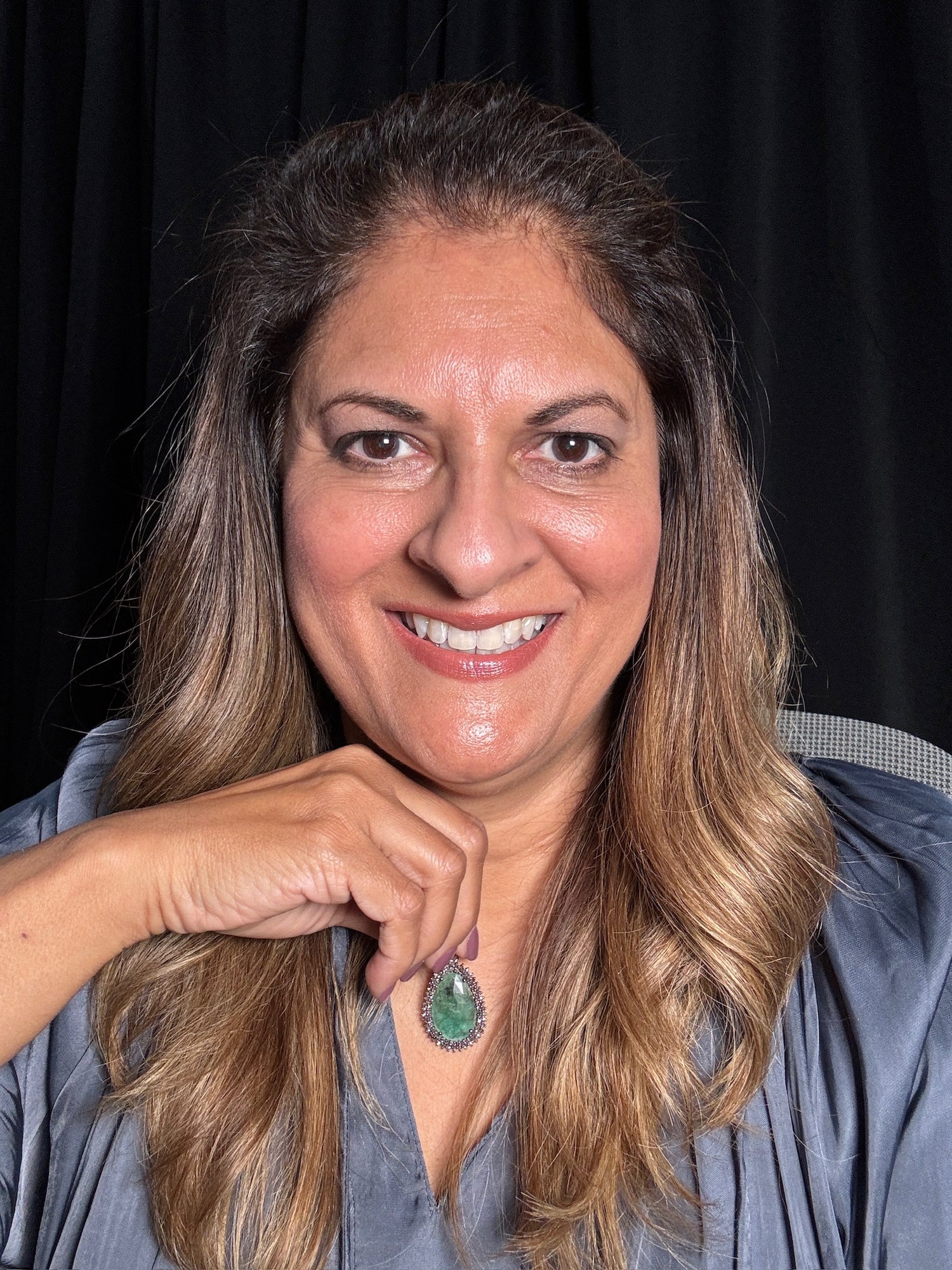 Smiling woman with long wavy brown hair wearing oval emerald pendant with diamond halo on sterling silver chain, photographed against black background