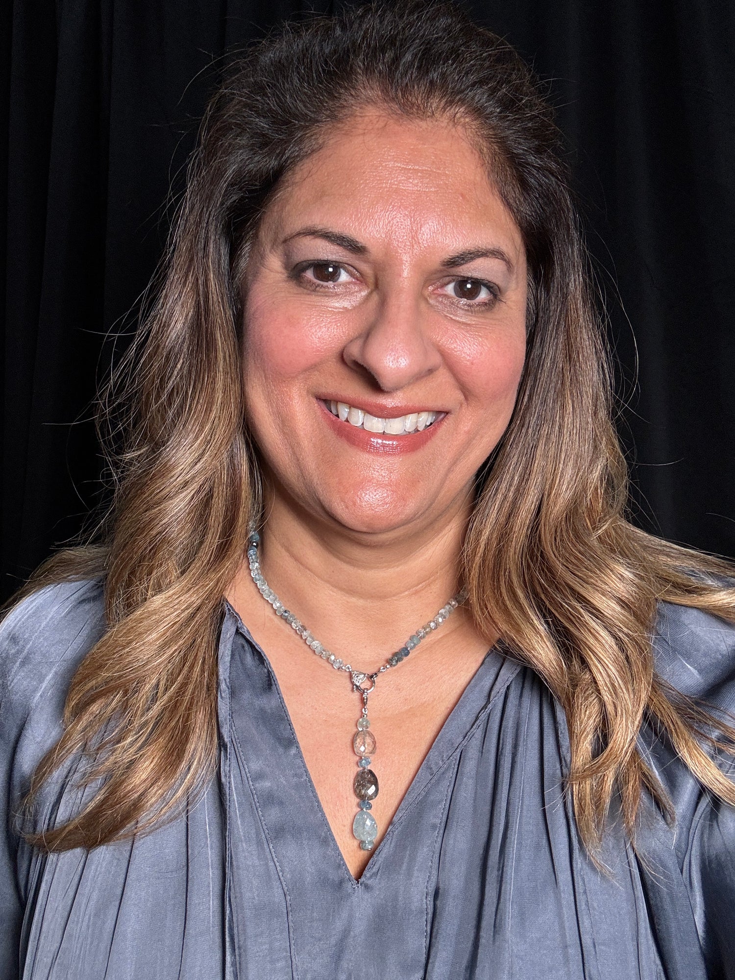 Woman with long wavy hair wearing gray blouse and aquamarine nugget necklace with silver chain against black background