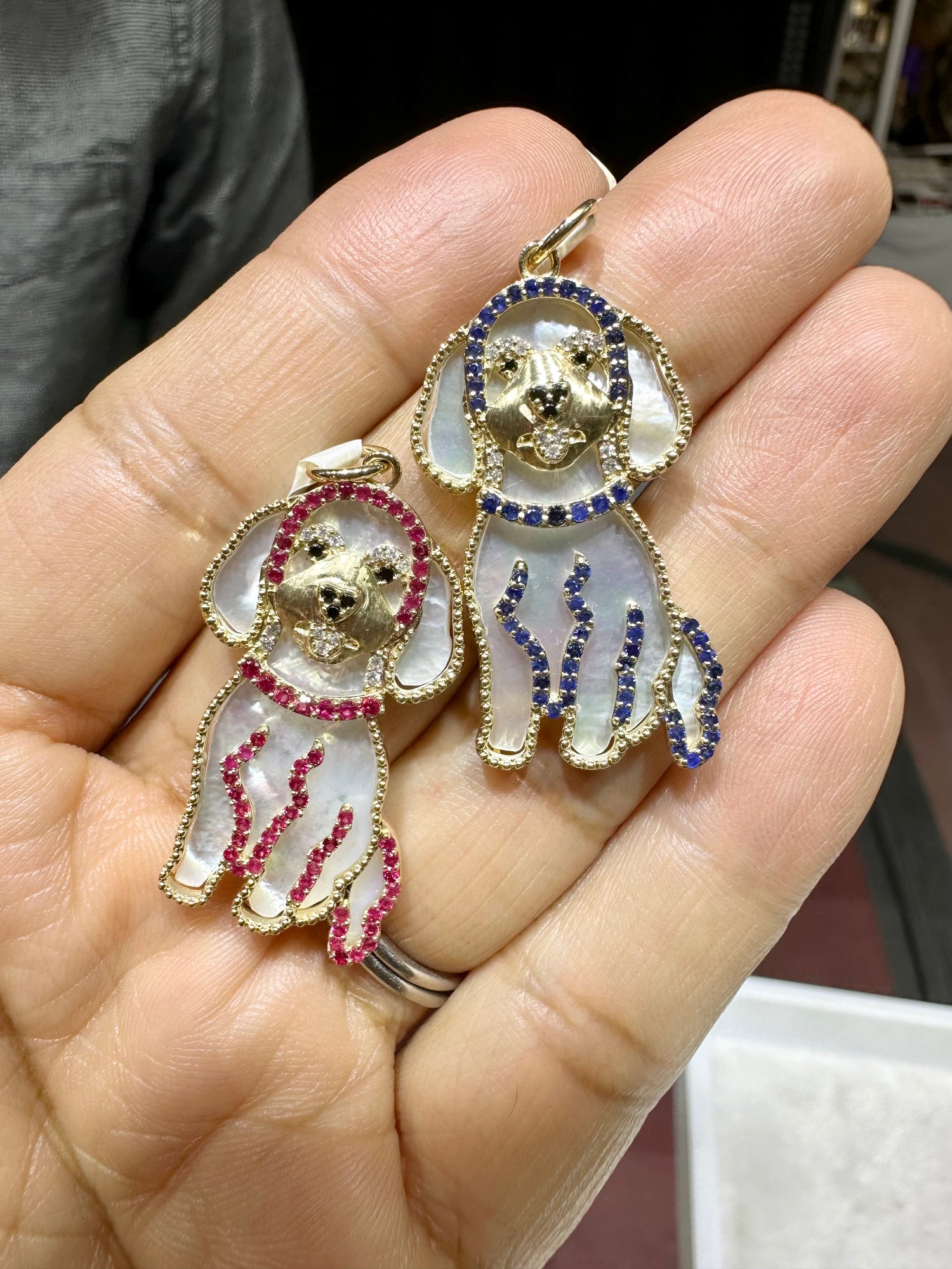 Two 14k gold Golden Doodle pendants held in palm - one with ruby and pink mother-of-pearl, one with sapphire and blue mother-of-pearl accents