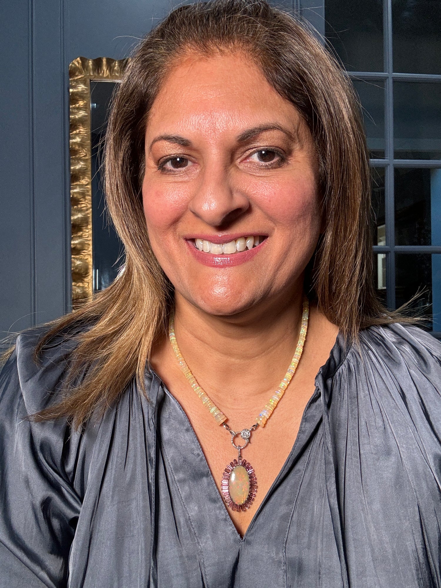 Smiling woman wearing the Ethiopian opal necklace with tourmaline-framed pendant against gray blouse, showing the elegant length and statement presence