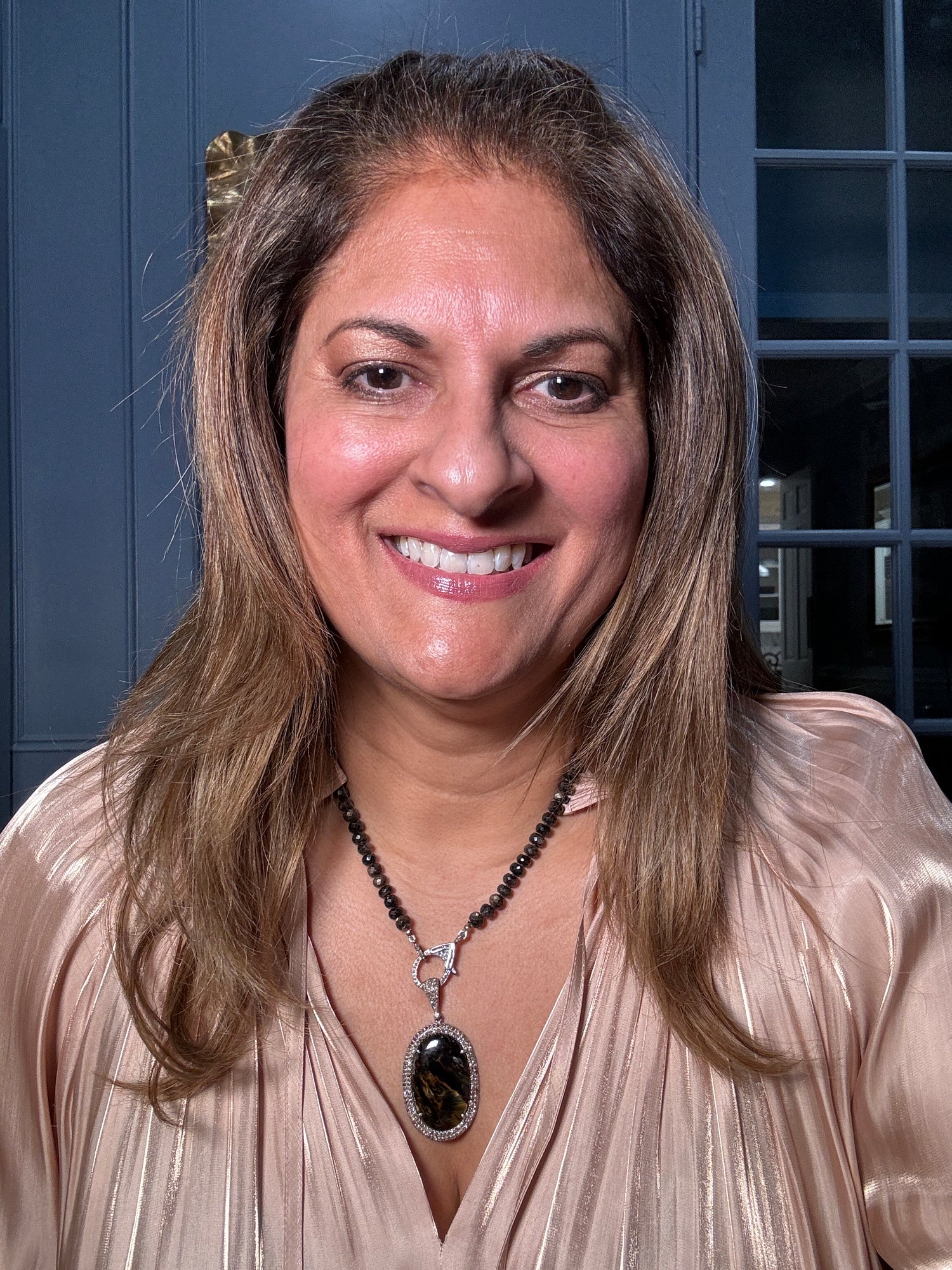 Woman wearing metallic gemstone necklace with dramatic pietersite pendant featuring golden-brown swirls and diamond halo against champagne blouse