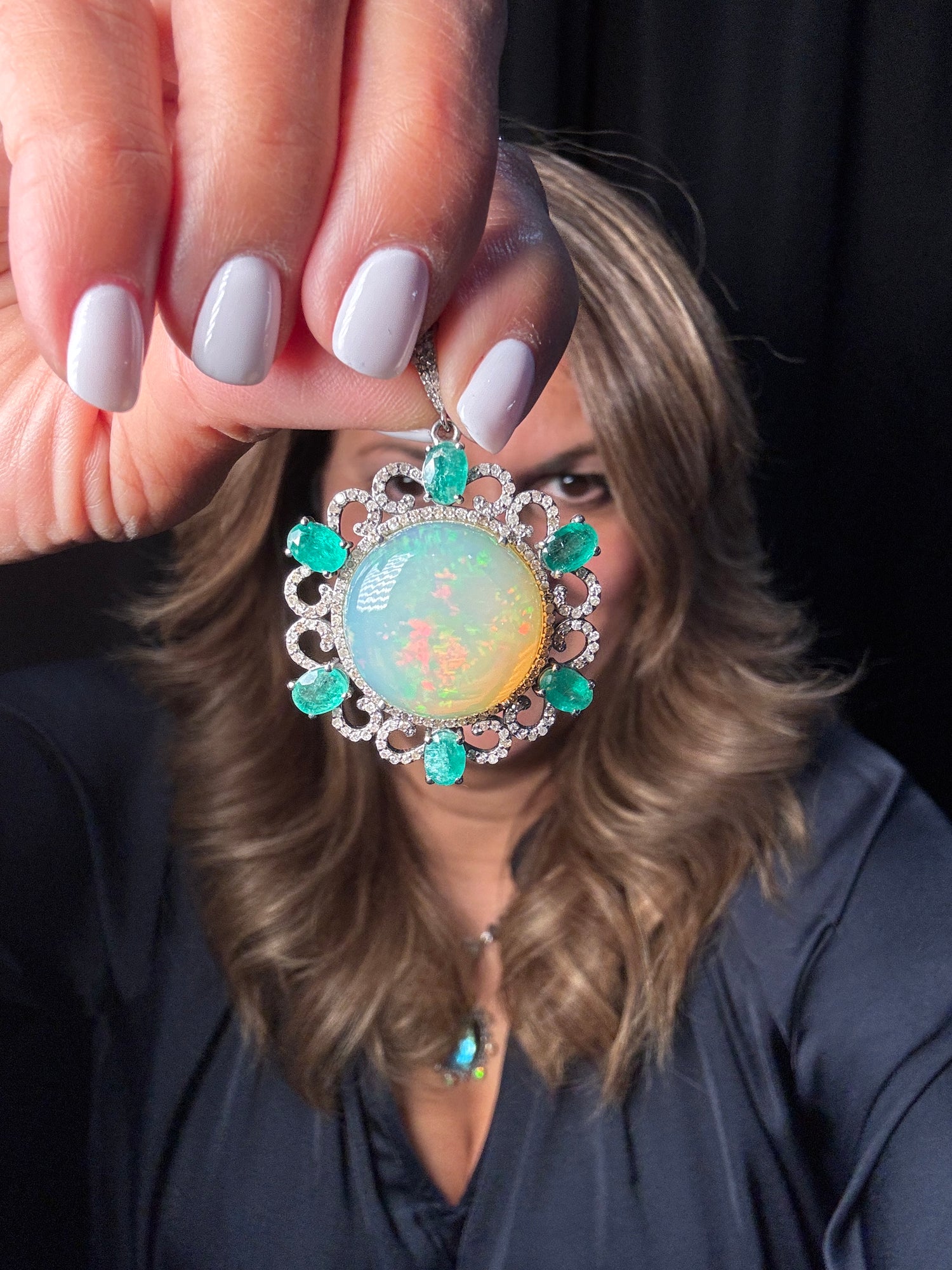 Hand holding ornate sterling silver pendant featuring large opal center stone surrounded by emeralds and diamonds in decorative floral setting