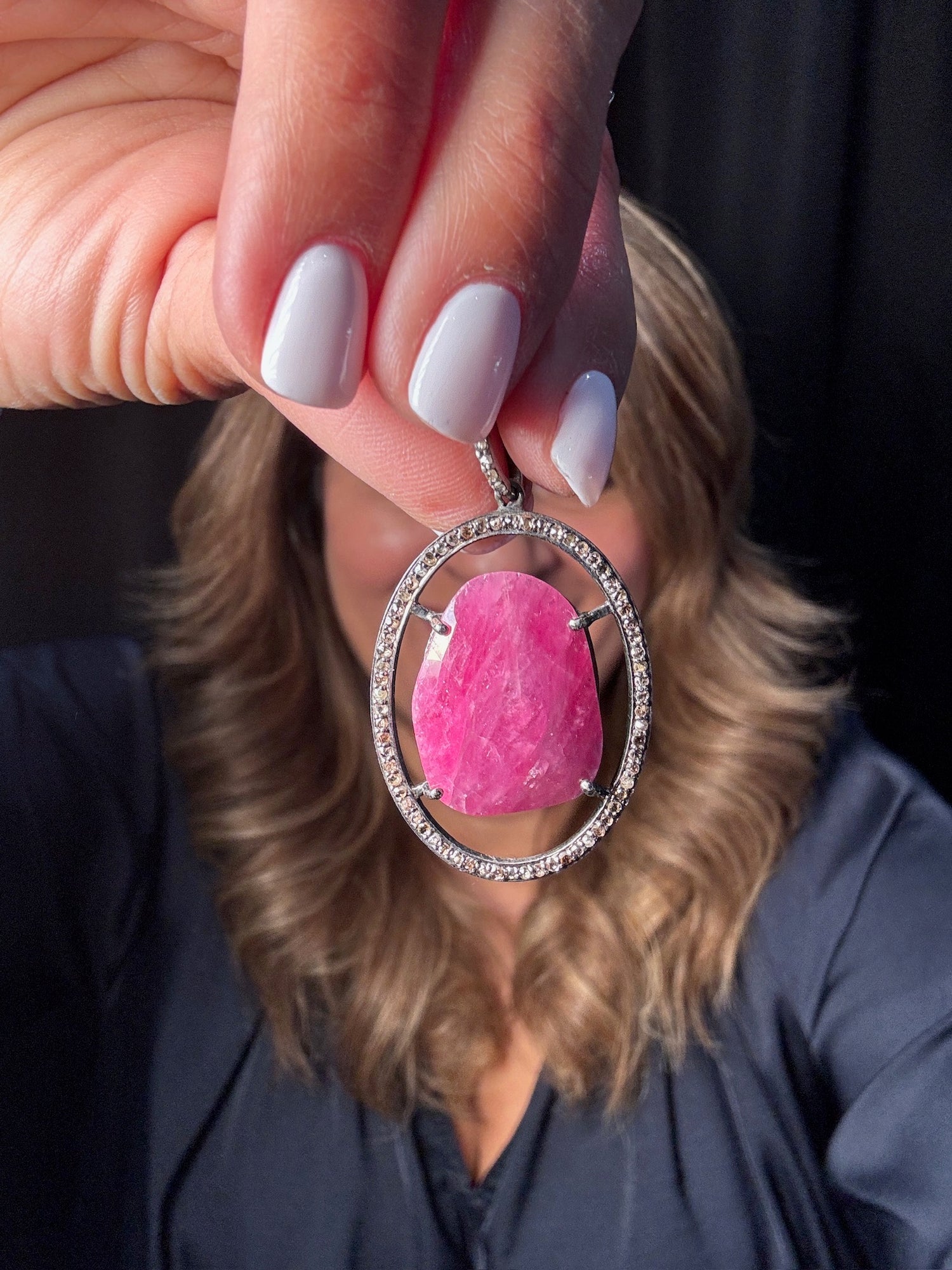 Hand holding large oval-cut pink ruby pendant with diamond-studded sterling silver frame against blonde-haired woman in navy shirt