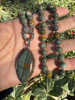 Hand holding 18-inch Rocky Butte jasper necklace outdoors in natural sunlight, showcasing varied green and amber tones in round beads and pendant