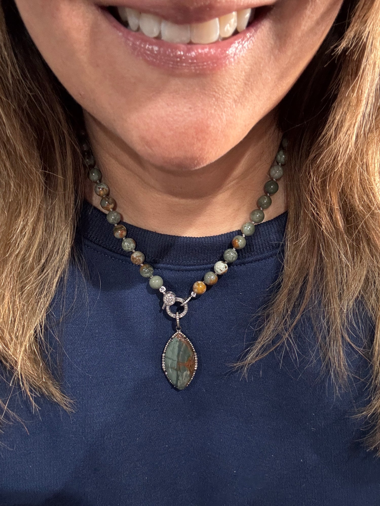 Smiling woman wearing Rocky Butte jasper necklace with green-brown beads and marquise pendant against navy blue top, showing elegant neckline styling