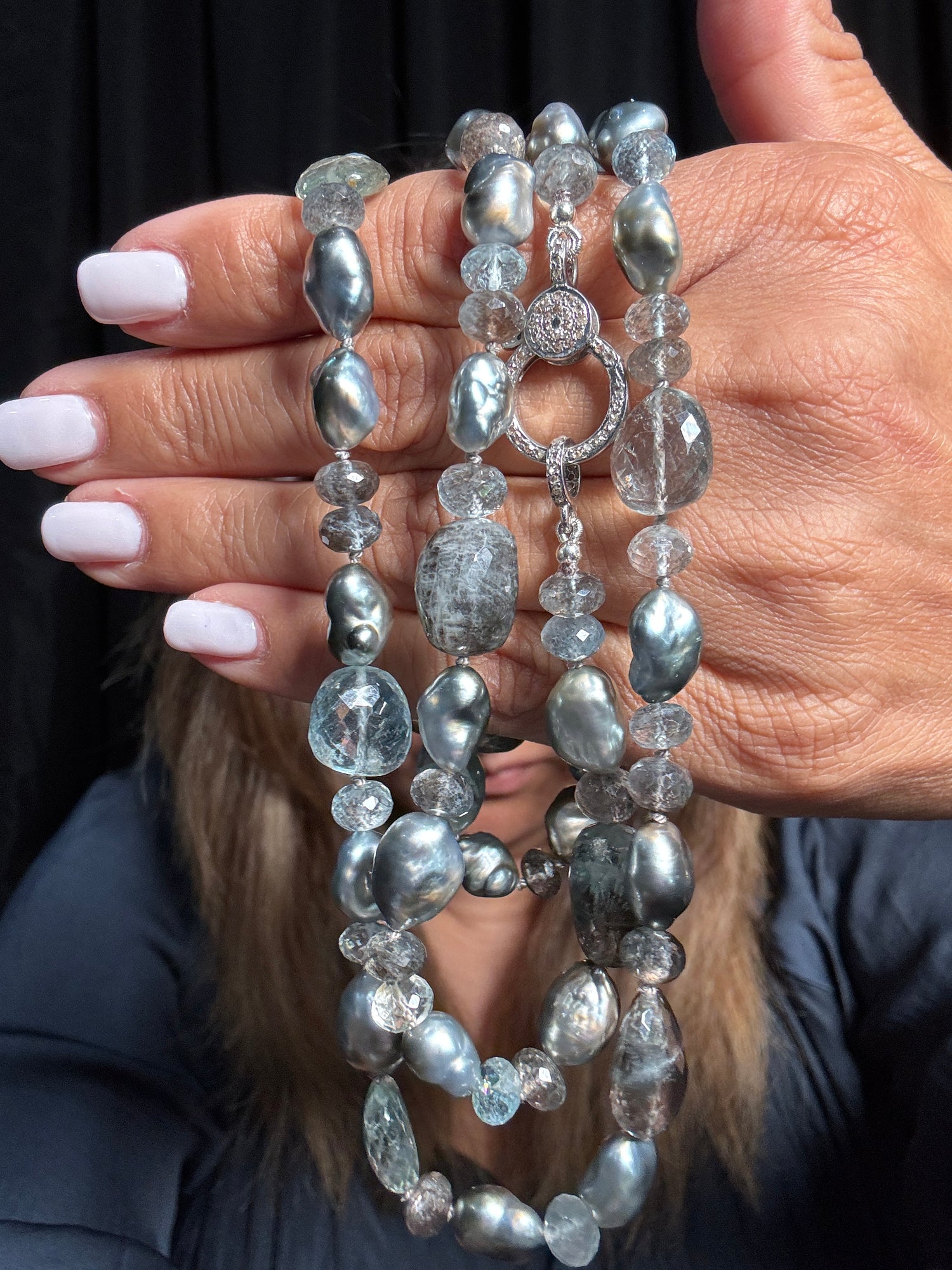 Elegant 36-inch necklace with blue-gray aquamarine gemstones and silver Tahitian keshi pearls featuring ornate diamond clasp, held gracefully in hands
