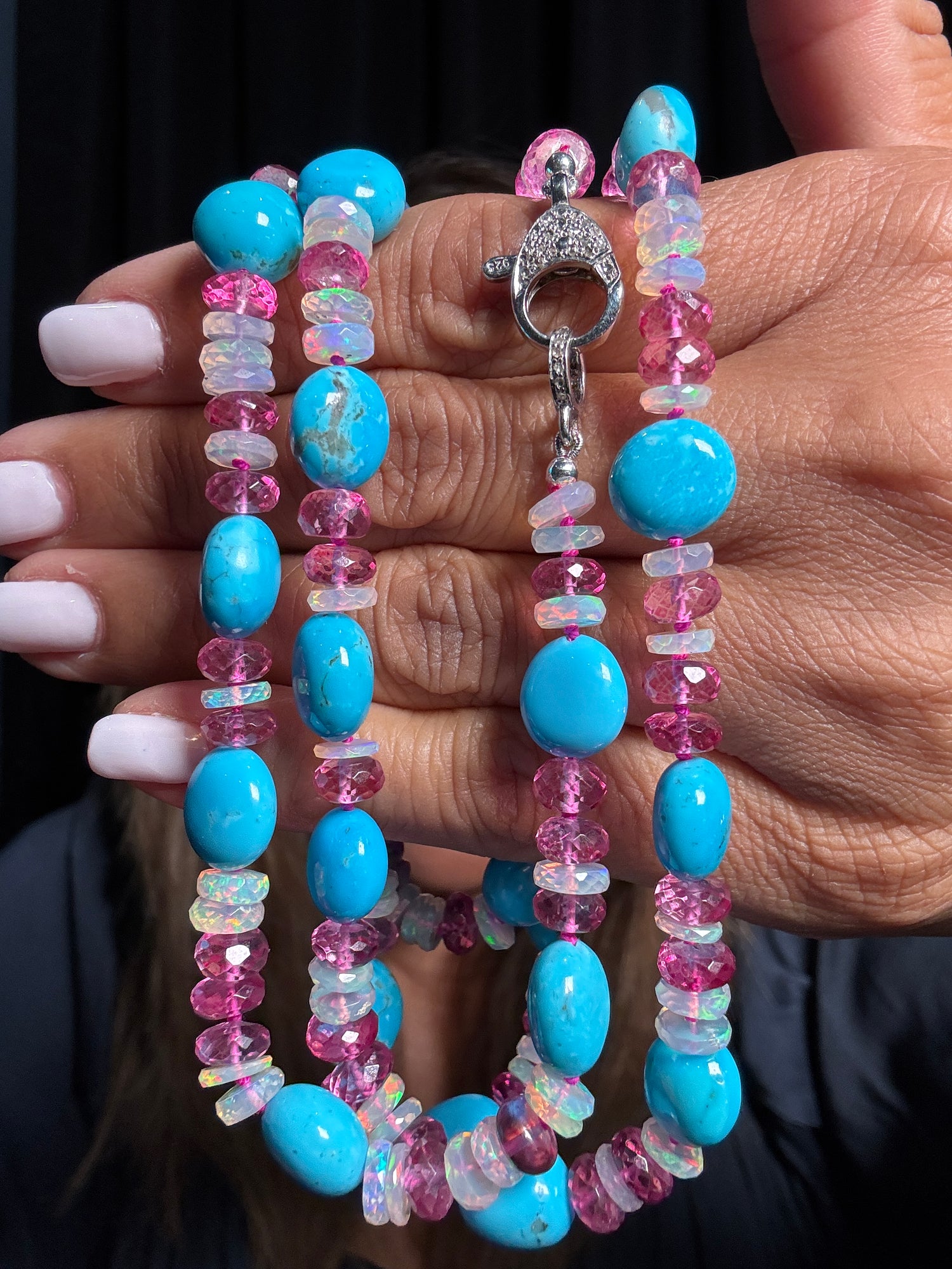Hands holding a 36-inch necklace featuring bright turquoise oval beads, pink tourmaline rondelles, and iridescent Ethiopian opal chips with silver diamond clasp