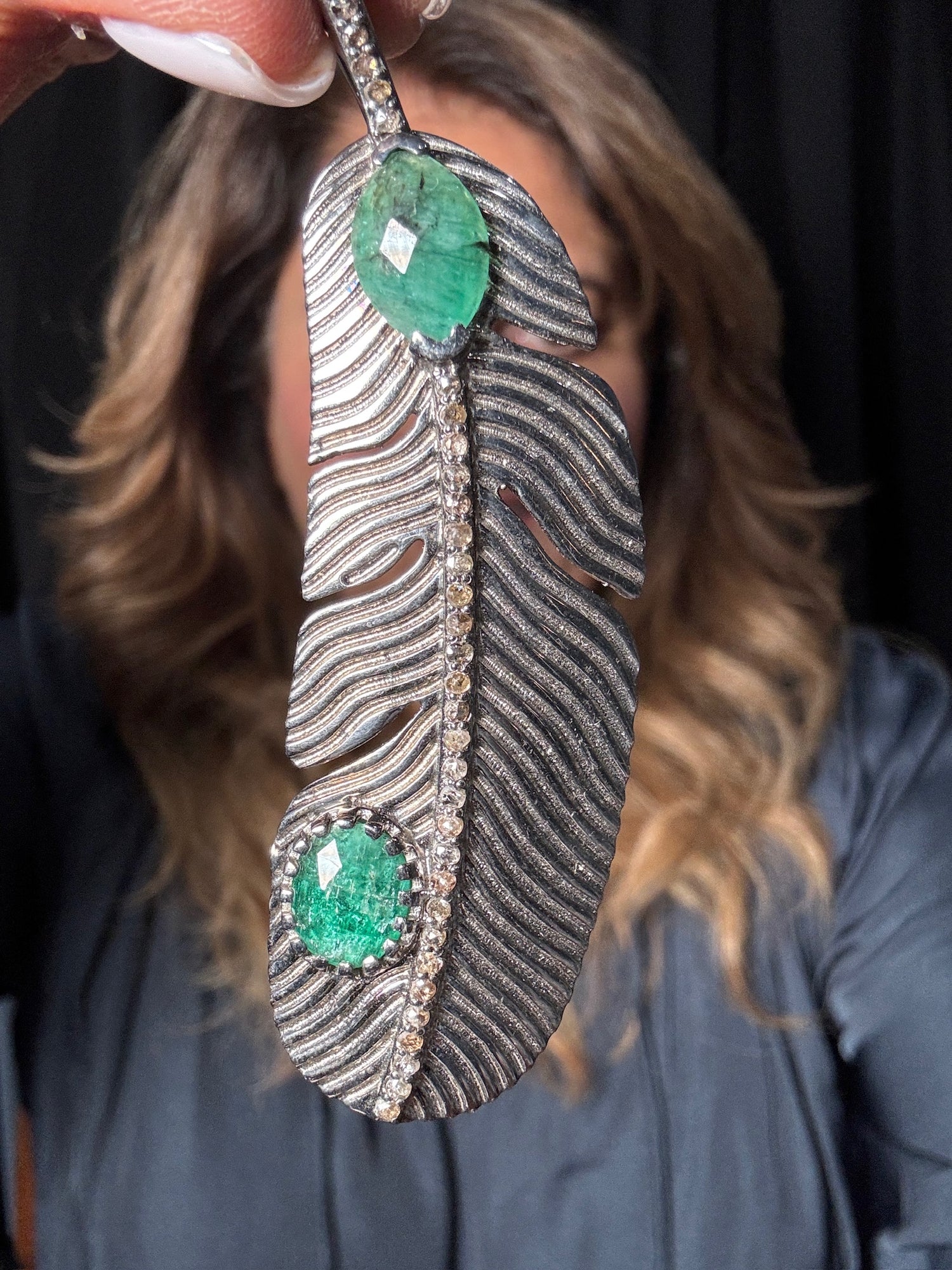 Close-up of hand holding sterling silver feather pendant with raw emeralds and diamonds, showing intricate textured metalwork and brilliant green gemstones