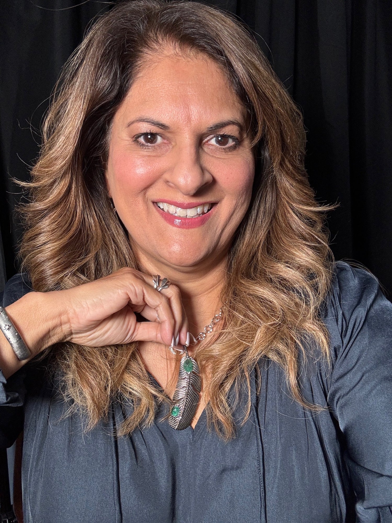 Smiling woman wearing sterling silver feather pendant with raw emeralds and diamonds on chain, displayed against dark background in portrait style