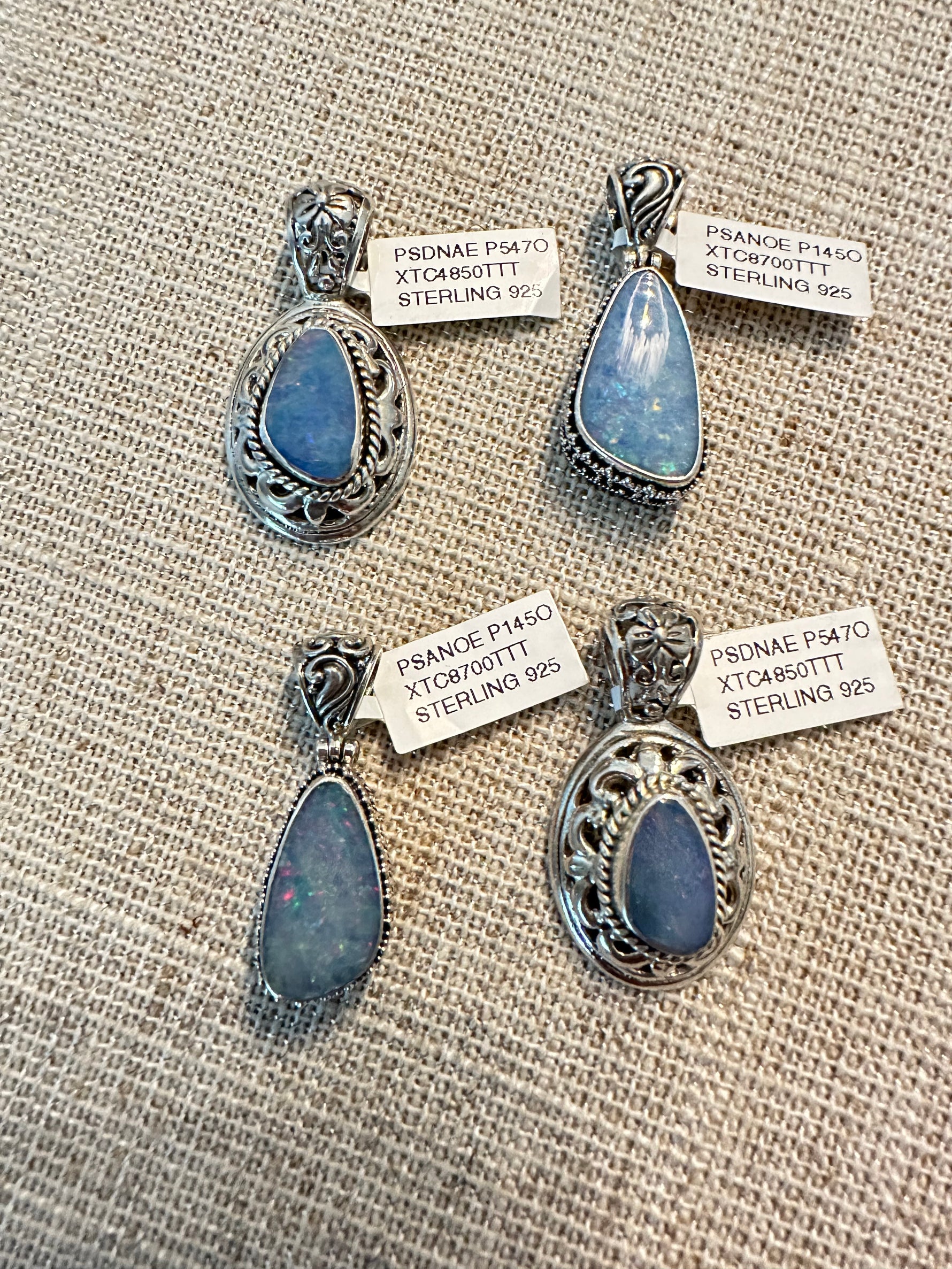 Four sterling silver Australian opal pendants displayed on natural burlap fabric, featuring blue opals in ornate and simple settings with price tags