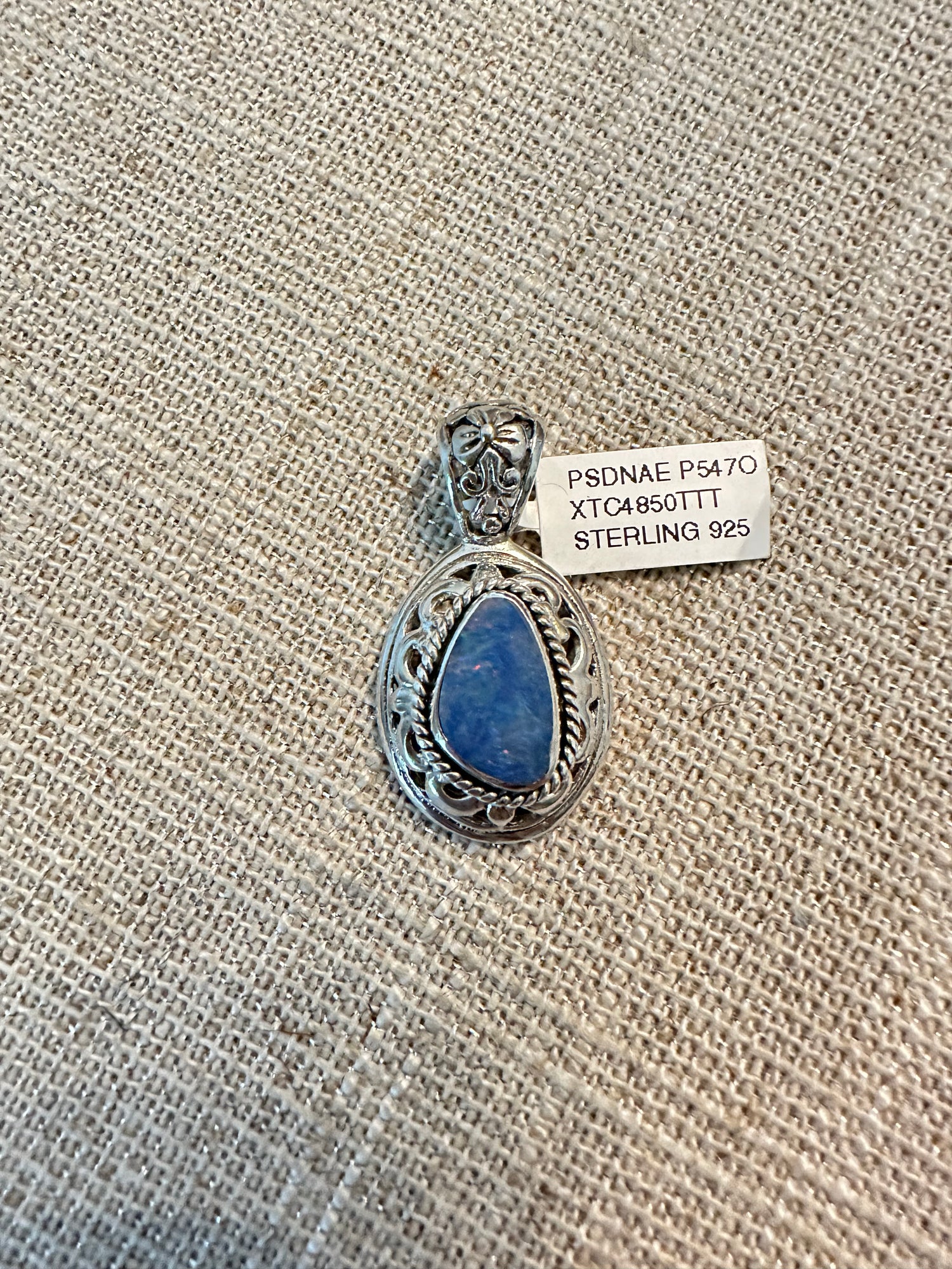 Ornate sterling silver pendant with oval blue Australian opal in decorative twisted rope border setting, photographed on textured burlap background