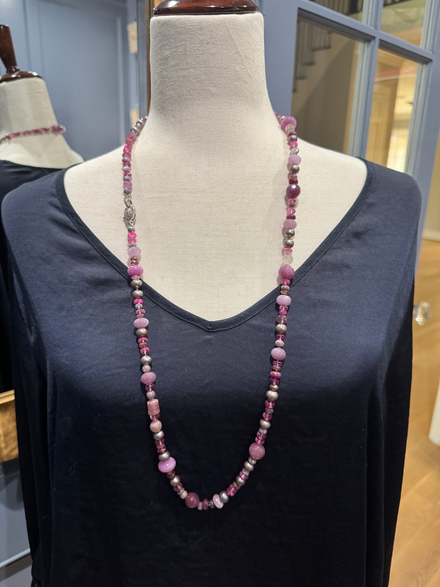 Long ruby kunzite and gray pearl necklace draped on cream fabric mannequin wearing black v-neck top in jewelry store display