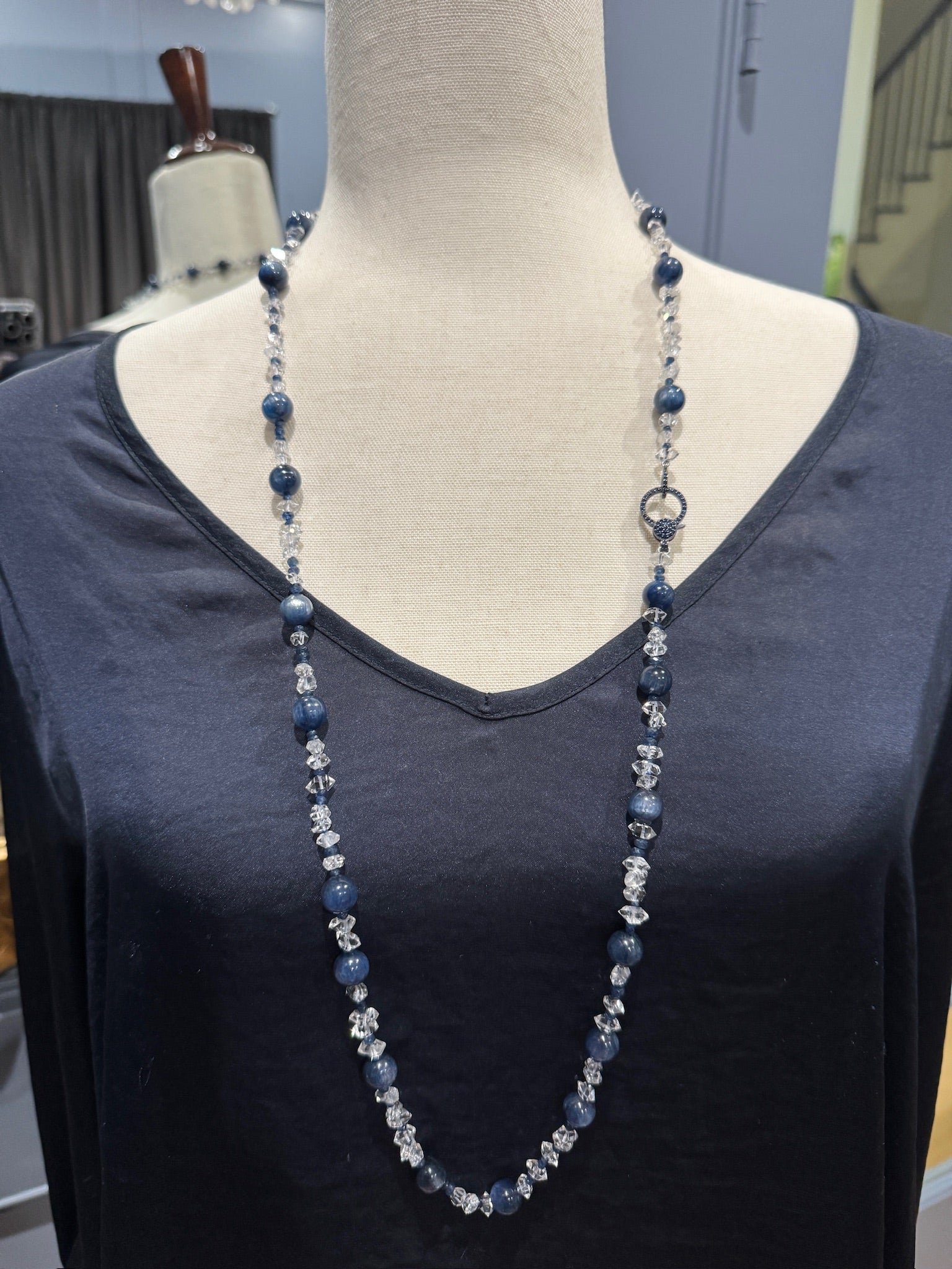 Long layering necklace with blue kyanite beads and sparkling clear Herkimer diamonds displayed on cream fabric mannequin wearing navy v-neck top.