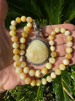 Hands cupping yellow opal bead necklace with large round yellow aquamarine pendant in diamond halo setting against green palm leaves