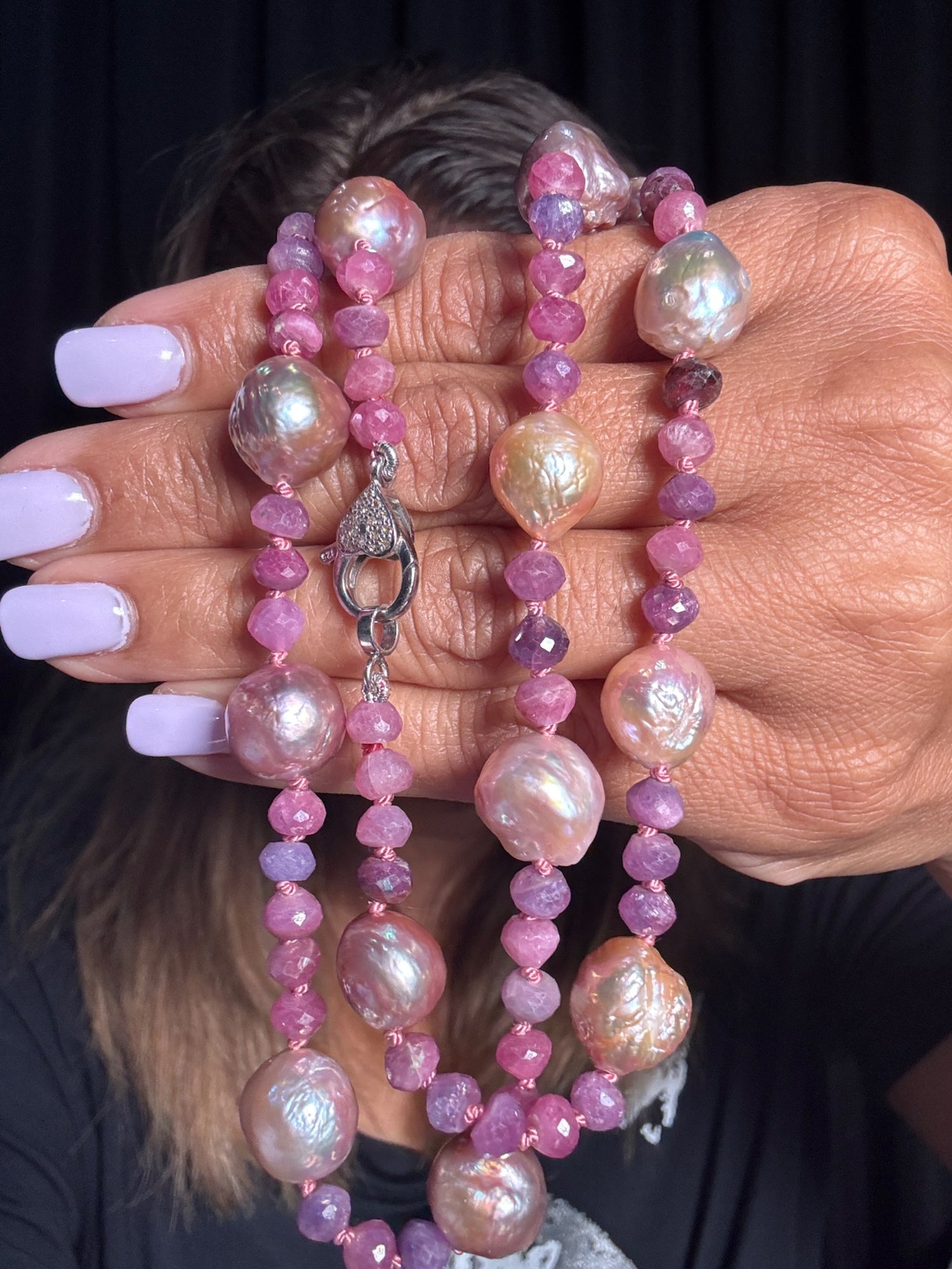 Hands holding a 23-25 inch necklace featuring lustrous pink Edison pearls and faceted ruby gemstones with sterling silver toggle clasp against dark background