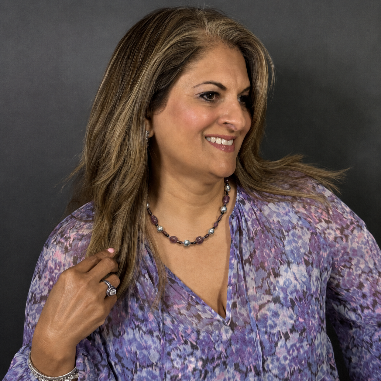 A smiling woman with shoulder-length brown hair wearing a purple and lavender floral print blouse, a beaded purple and silver necklace, and a silver ring, posing against a dark gray studio background.