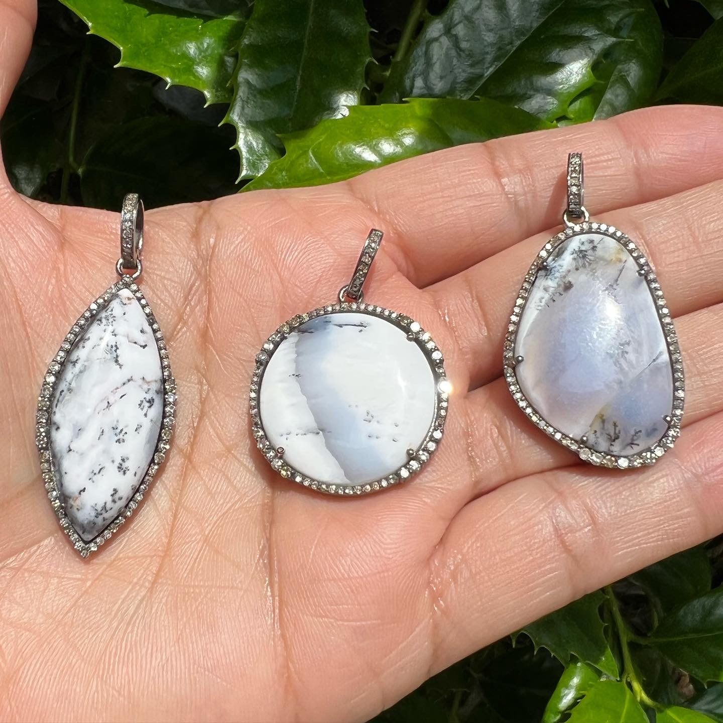 Three dendrite opal pendants with diamond halos held in palm, showing white opal with black dendritic inclusions in sterling silver settings