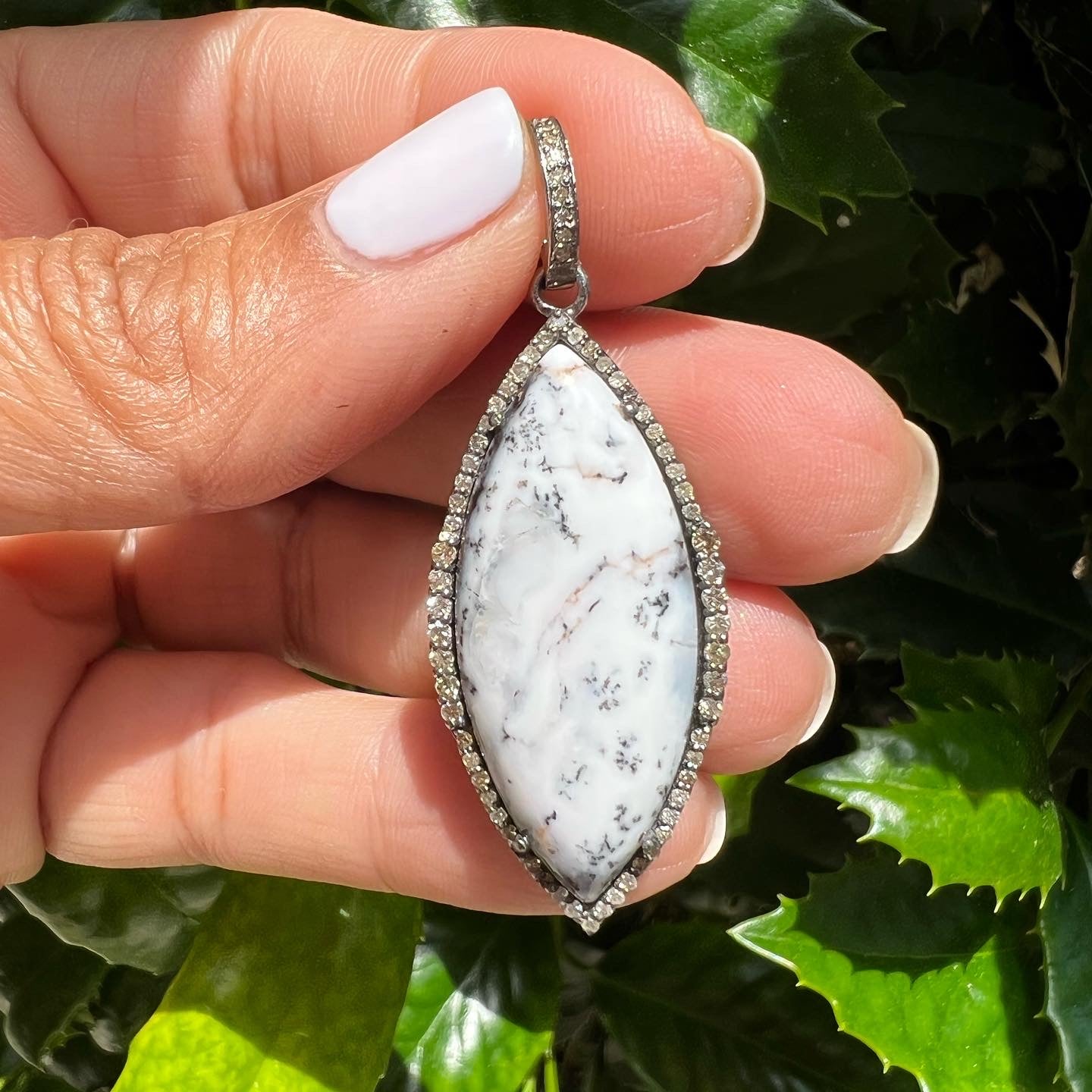 Marquise-shaped dendrite opal pendant with diamond halo held between fingers, displaying white opal stone with black tree-like mineral inclusions